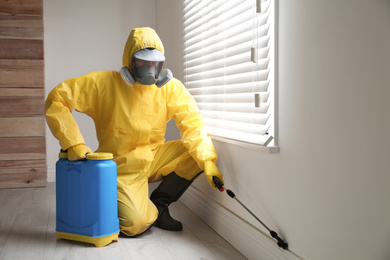 Photo of Pest control worker in protective suit spraying pesticide near window indoors