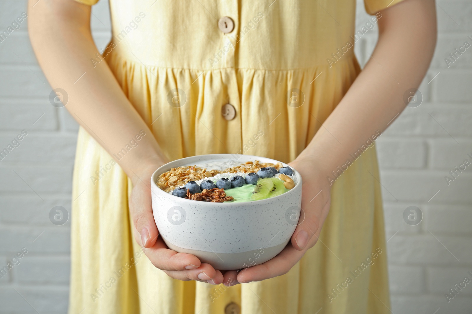 Photo of Woman holding tasty smoothie bowl with fresh kiwi fruit, berries and granola near white brick wall, closeup