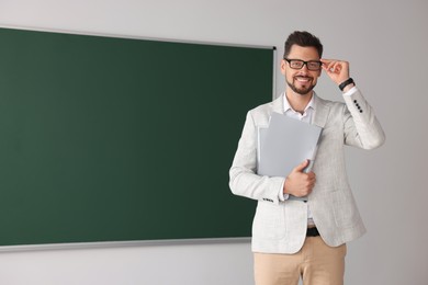 Photo of Happy teacher with glasses and stationery at blackboard in classroom. Space for text