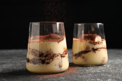 Photo of Adding cocoa powder into glass with delicious tiramisu at grey textured table, closeup