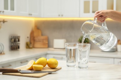 Photo of Woman pouring water from jug into glass at white table in kitchen, closeup