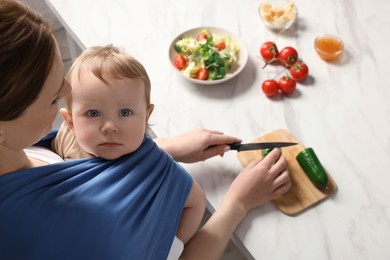 Mother cutting cucumber while holding her child in sling (baby carrier) indoors, above view