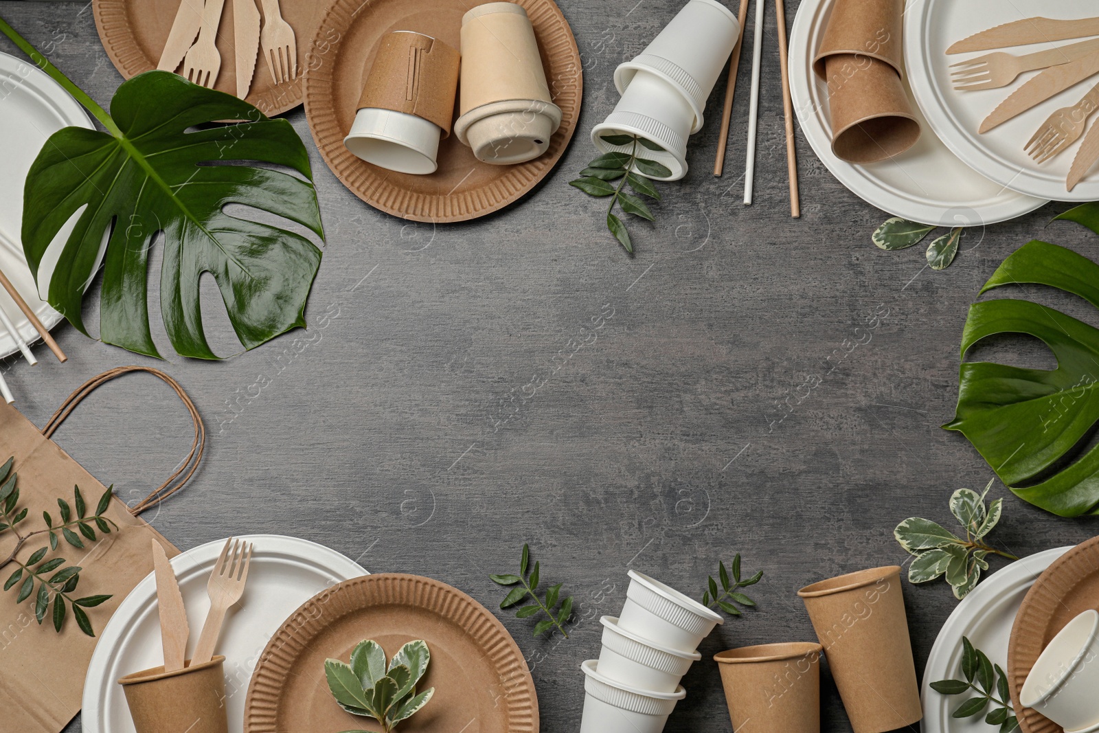 Photo of Frame of disposable tableware and different green leaves on grey background, flat lay. Space for text