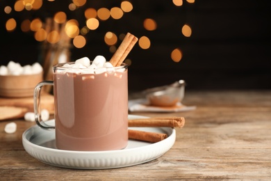 Photo of Glass cup of hot cocoa with aromatic cinnamon and marshmallows on wooden table against blurred lights. Space for text