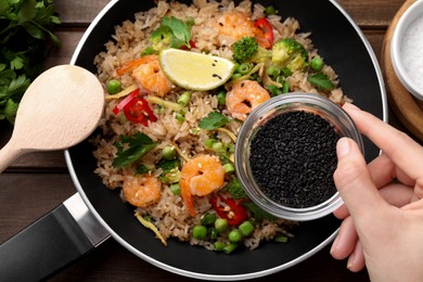 Woman adding sesame seeds to rice with shrimps and vegetables at wooden table, top view