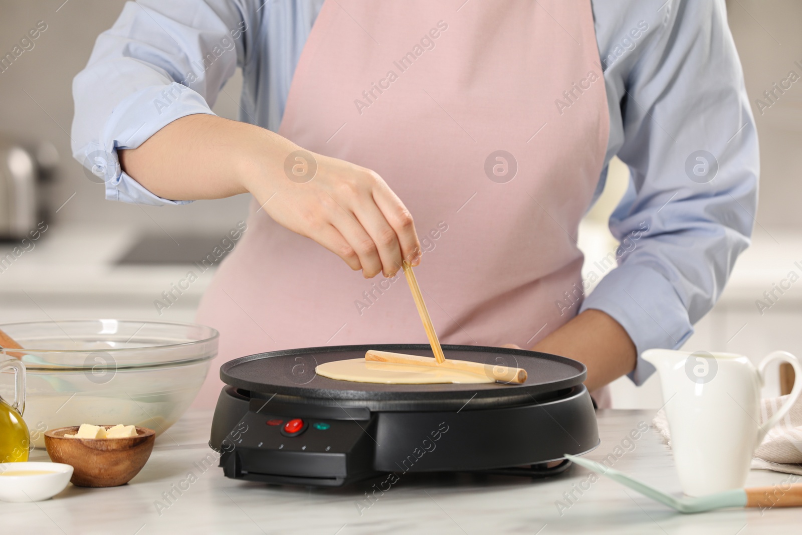 Photo of Woman cooking delicious crepe on electric pancake maker at white marble table in kitchen, closeup