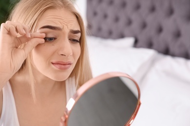 Young woman with eyelash loss problem looking in mirror indoors