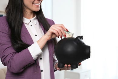 Businesswoman putting money into piggy bank indoors, closeup