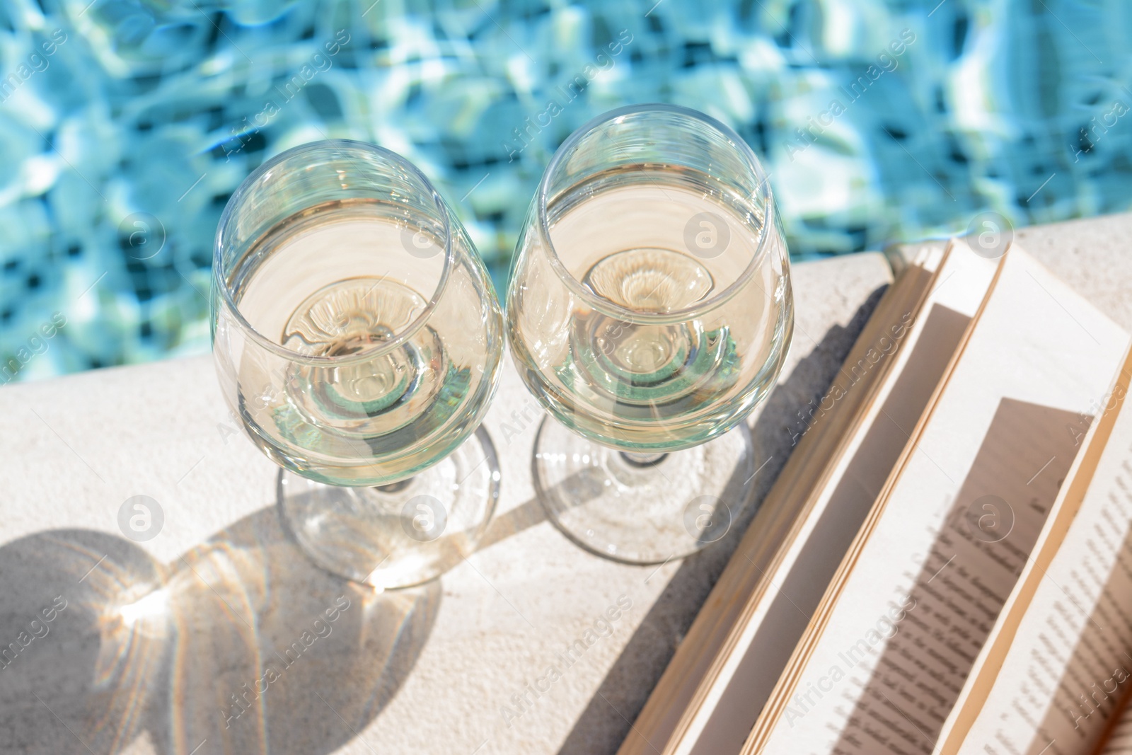 Photo of Glasses of tasty wine and open book on swimming pool edge, closeup