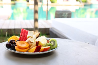 Plate with fresh fruits on table near sun lounger, space for text. Luxury resort with outdoor swimming pool