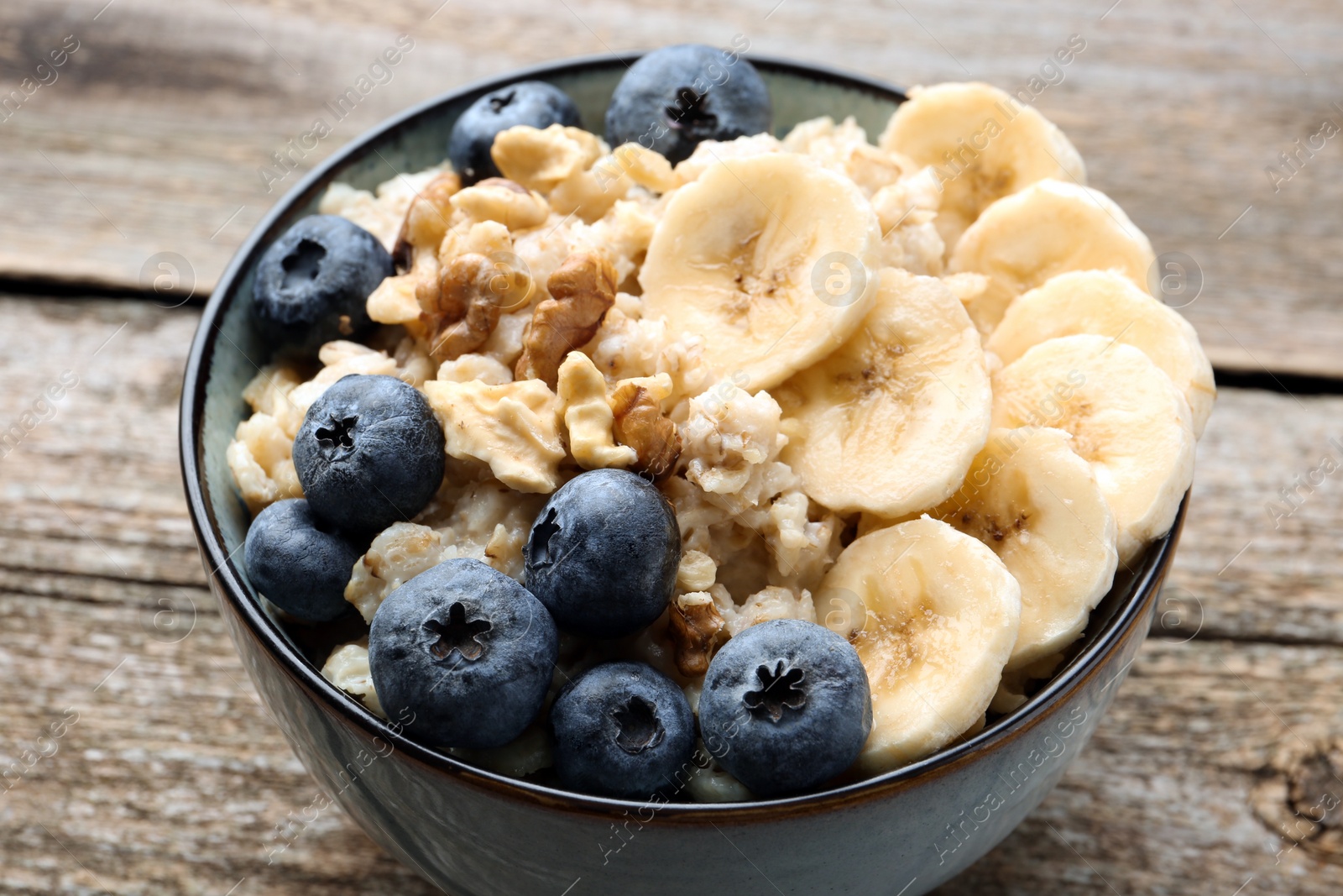 Photo of Tasty oatmeal with banana, blueberries and walnuts served in bowl on wooden table, closeup