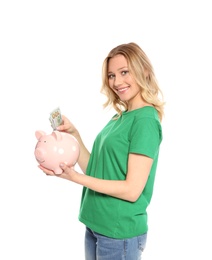 Beautiful young woman putting money into piggy bank on white background