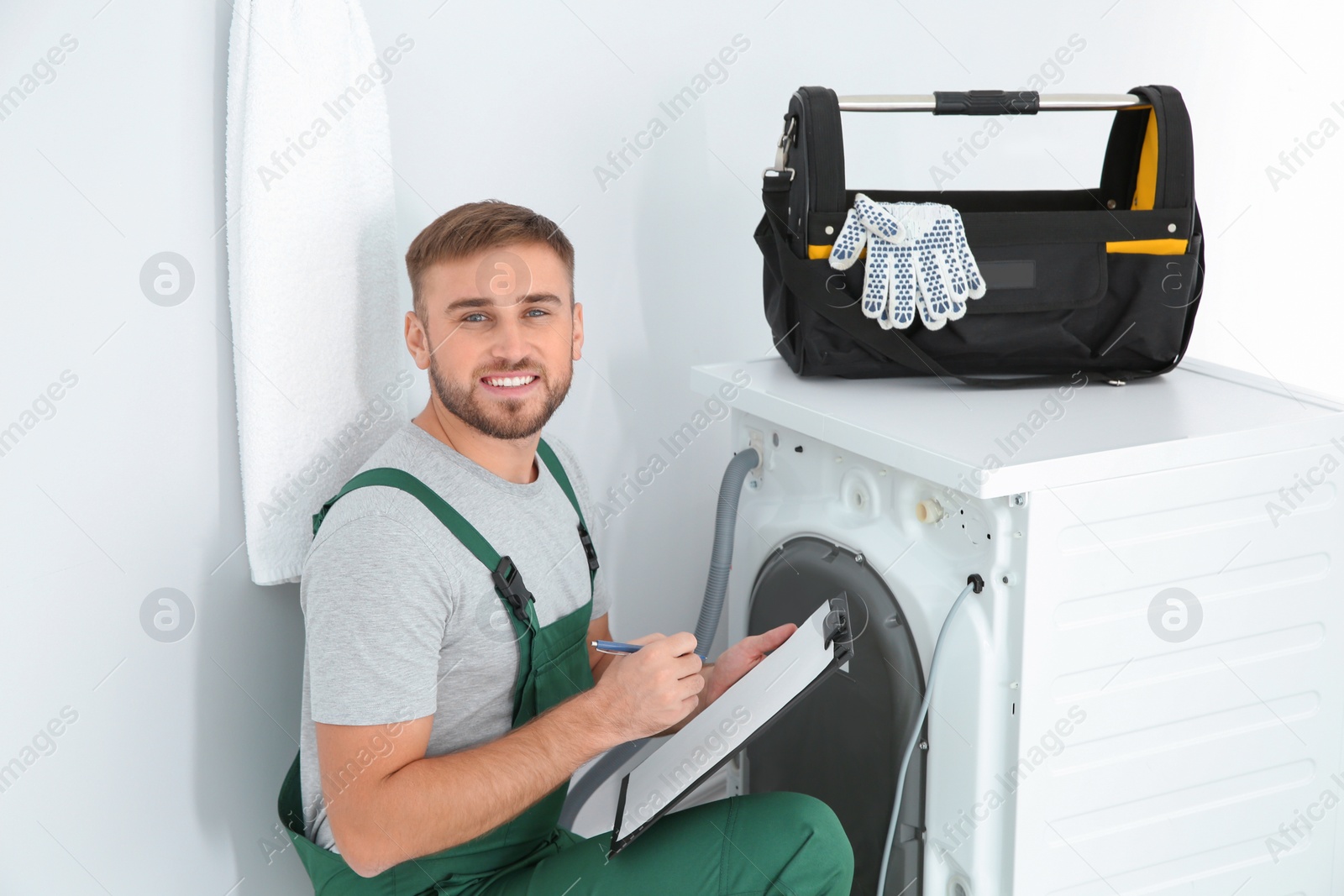 Photo of Young plumber with clipboard examining washing machine in bathroom