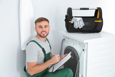 Young plumber with clipboard examining washing machine in bathroom