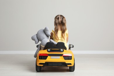 Cute little girl with toy elephant driving children's car near grey wall indoors, back view