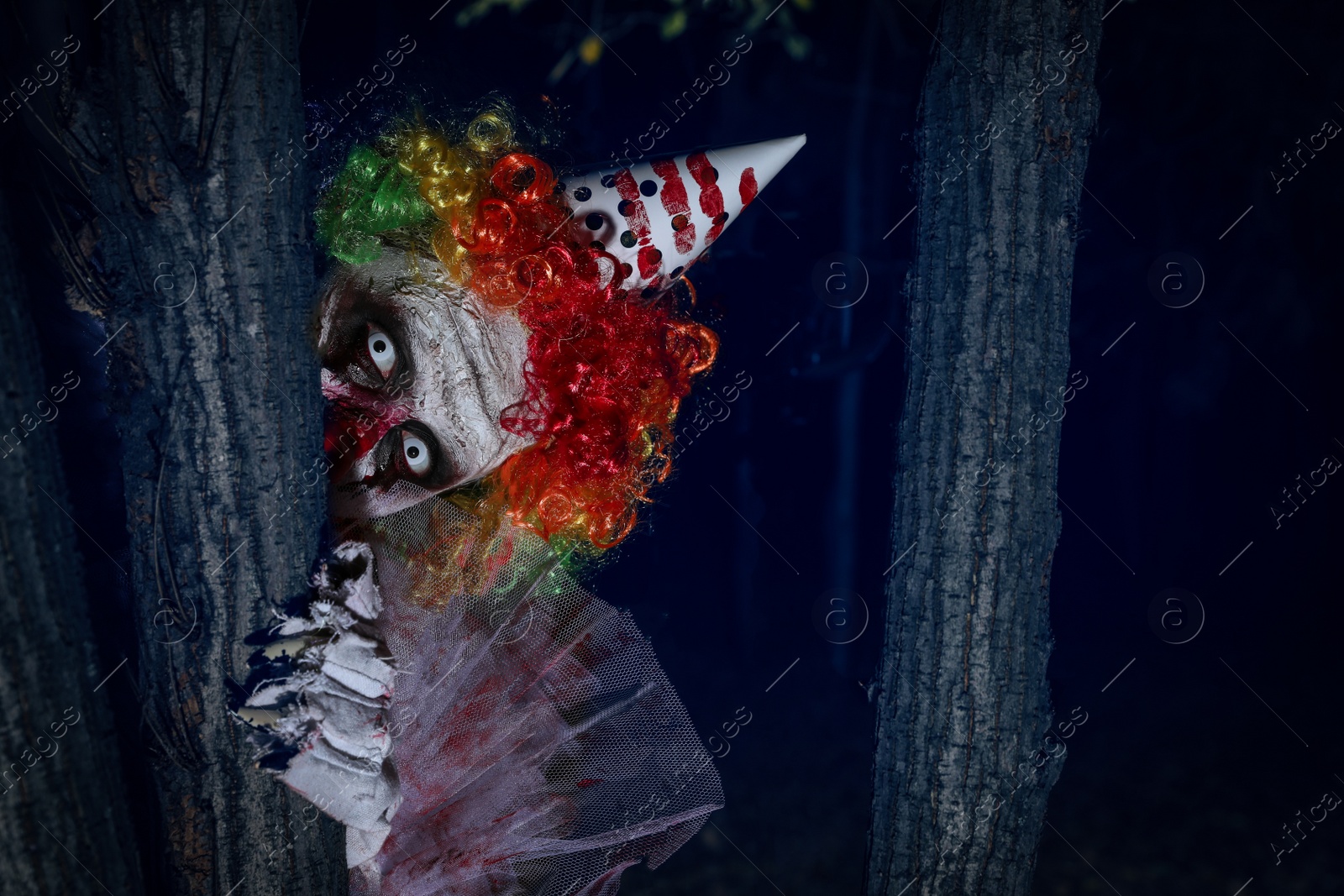 Photo of Terrifying clown hiding behind trees outdoors at night. Halloween party costume