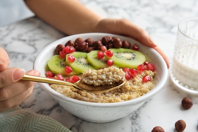 Photo of Woman eating quinoa porridge with hazelnuts, kiwi and pomegranate seeds at marble table, closeup