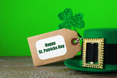 Green leprechaun hat, tag and clover leaf on wooden table against green background, closeup. St. Patrick's Day celebration