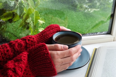 Photo of Woman with cup of hot drink near window on rainy day, closeup