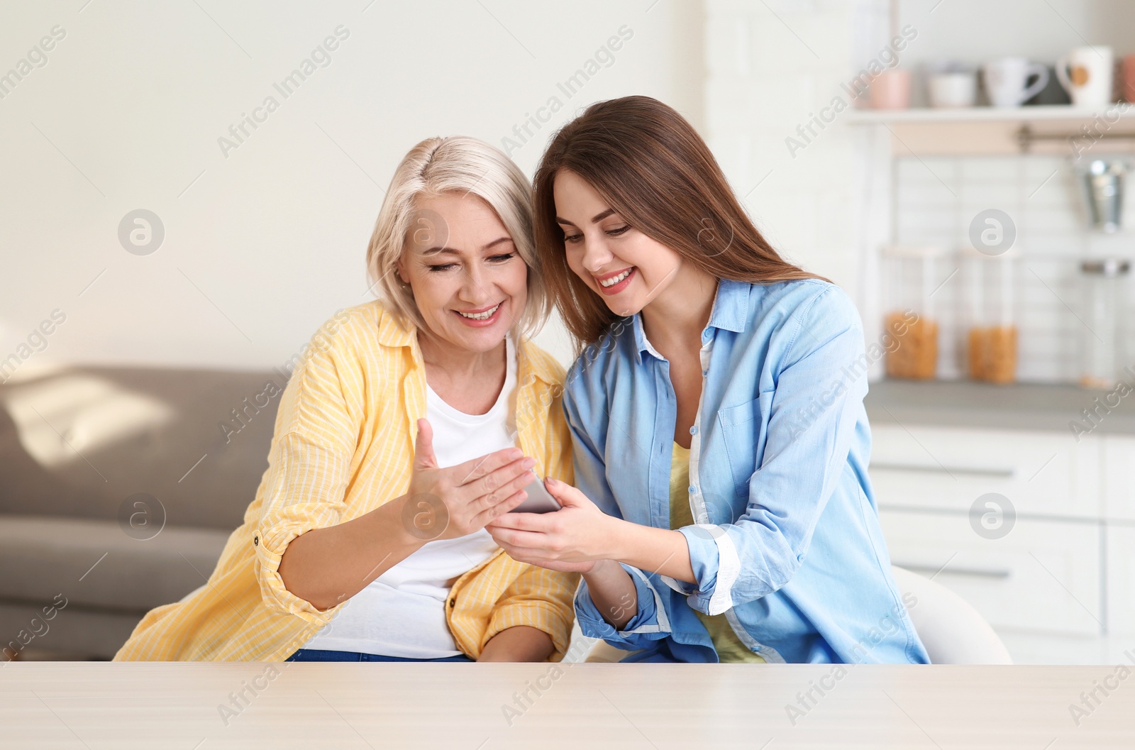 Photo of Portrait of young woman and her mature mother with mobile phone at table indoors