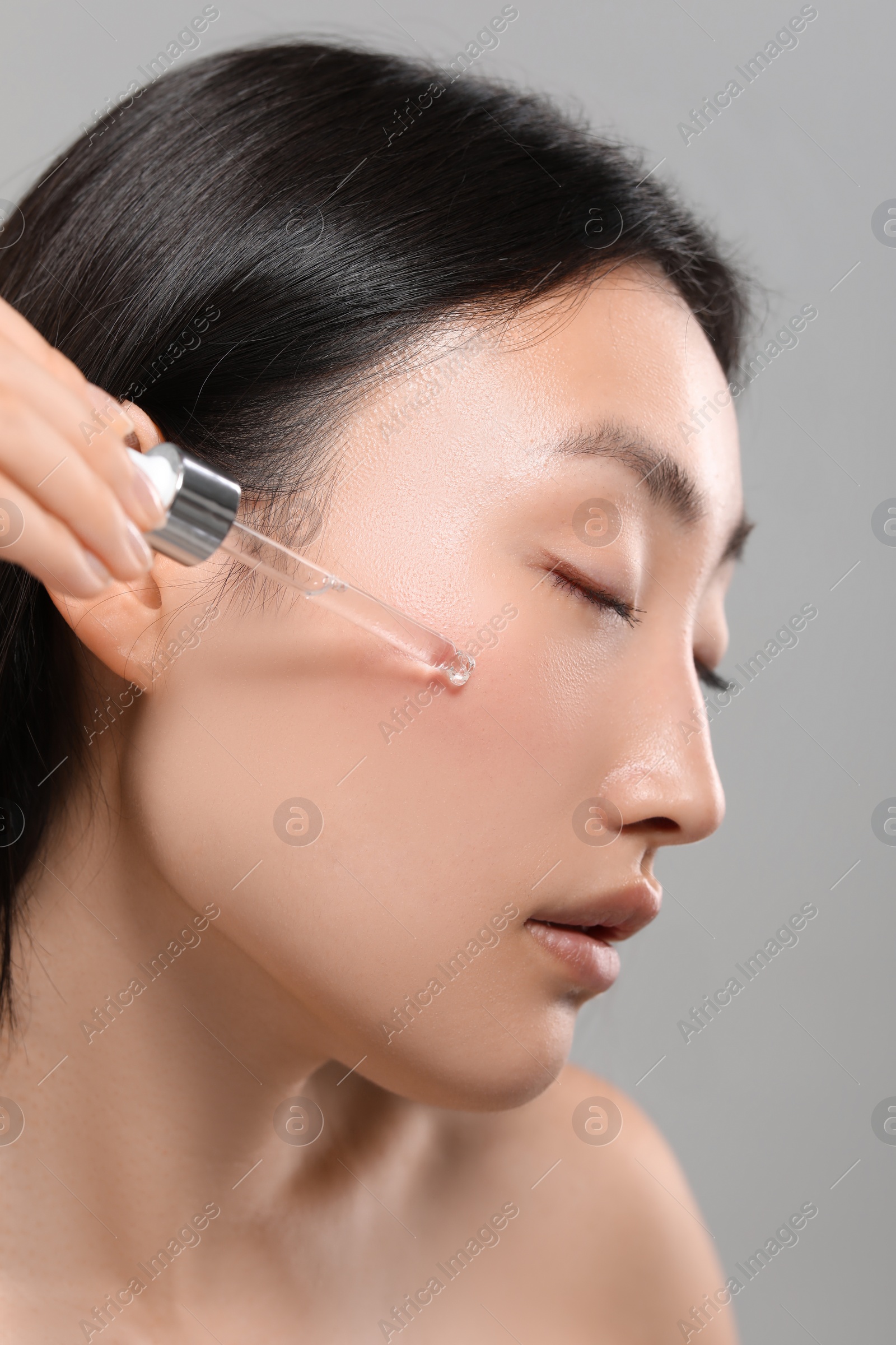 Photo of Beautiful young woman applying cosmetic serum onto her face on grey background, closeup