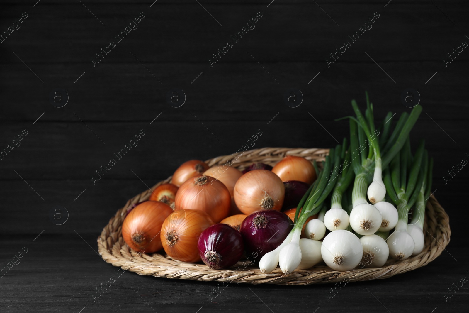 Photo of Wicker mat with different kinds of onions on black wooden table