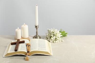 Photo of Church candles, wooden cross, rosary beads, Bible and flowers on light table. Space for text