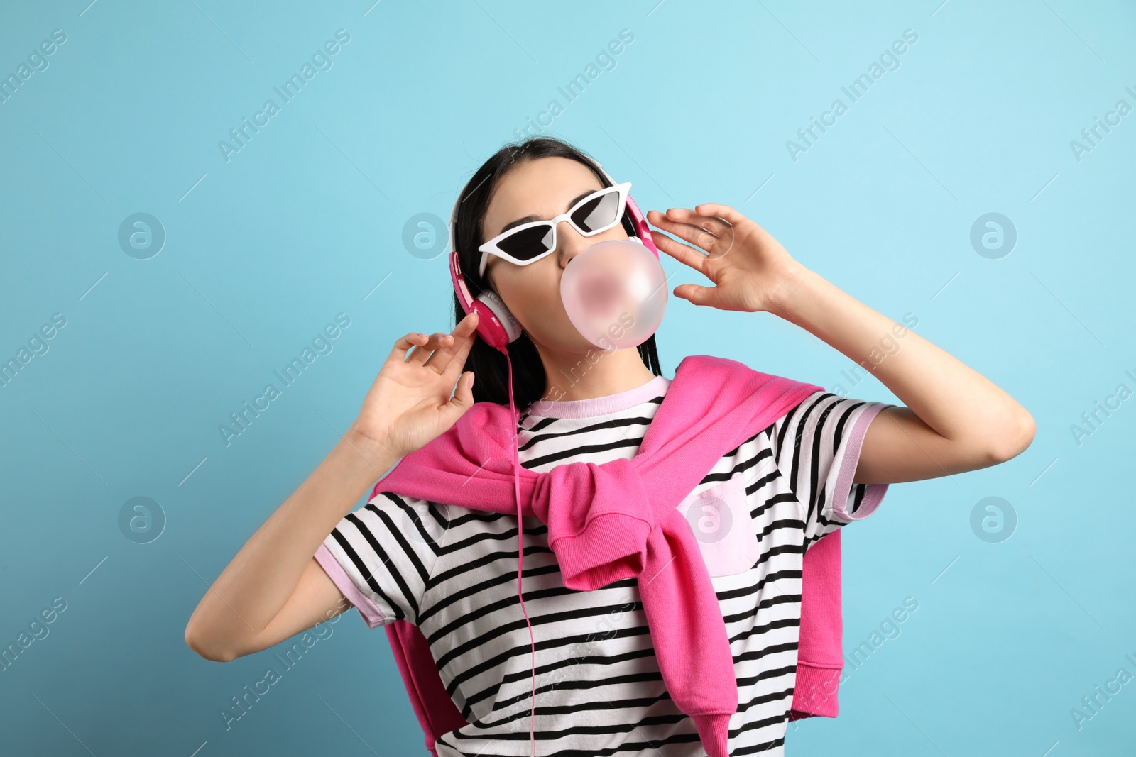 Photo of Fashionable young woman with headphones blowing bubblegum on light blue background