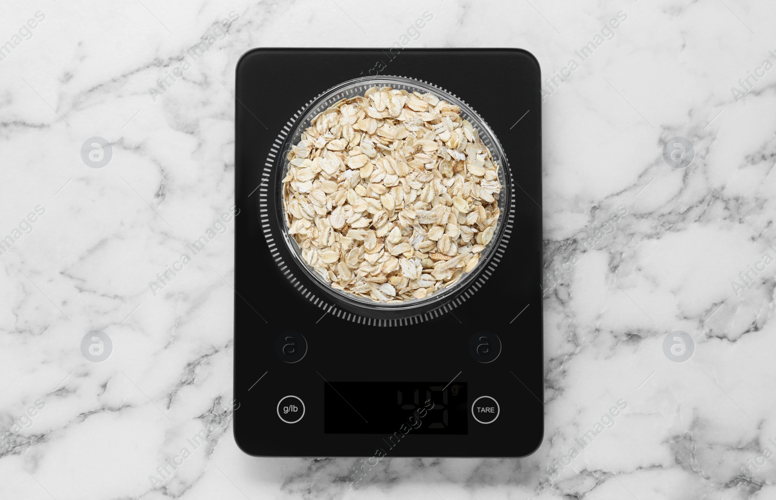 Photo of Digital kitchen scale with oat flakes on white marble table, top view