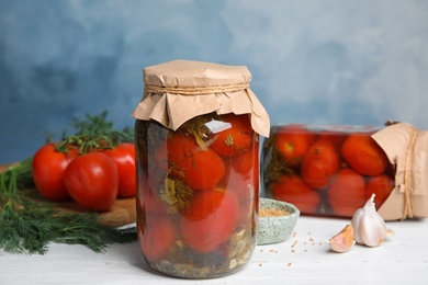 Photo of Pickled tomatoes in glass jars and products on white wooden table against blue background