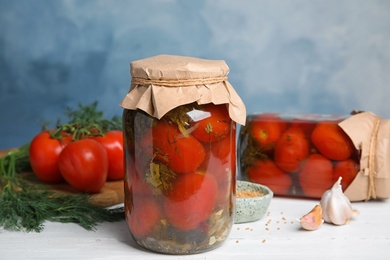 Pickled tomatoes in glass jars and products on white wooden table against blue background