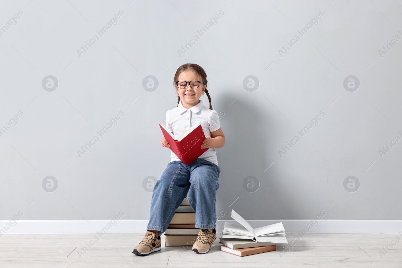 Photo of Cute little girl in glasses sitting on stack of books near light grey wall