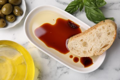 Bowl of organic balsamic vinegar with oil served with bread slice, basil and olives on white marble table, flat lay