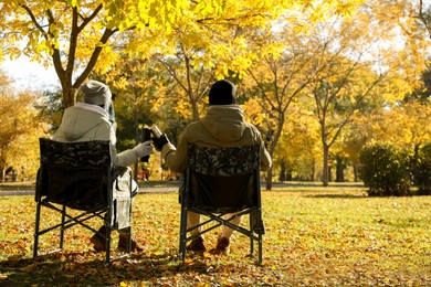 Couple with thermoses sitting in camping chairs outdoors on autumn sunny day, back view