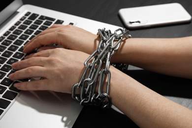 Woman with chained hands using laptop on black background, closeup. Loneliness concept