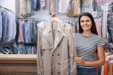 Photo of Dry-cleaning service. Happy woman holding hanger with coat in plastic bag indoors, space for text