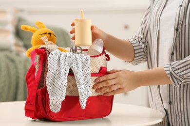 Photo of Woman putting sippy cup into mother`s bag at white table indoors, closeup