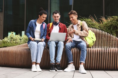 Photo of Happy young students studying with laptop on bench outdoors