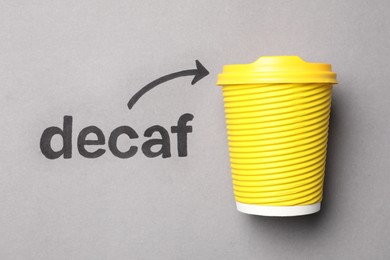 Word Decaf and arrow pointing at takeaway paper coffee cup on light grey background, top view