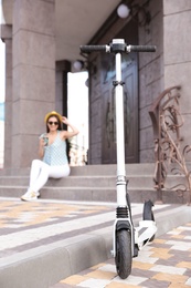 Photo of Young woman sitting near building outdoors, focus on electric kick scooter