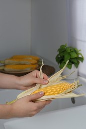 Photo of Woman husking corn cob in kitchen, closeup