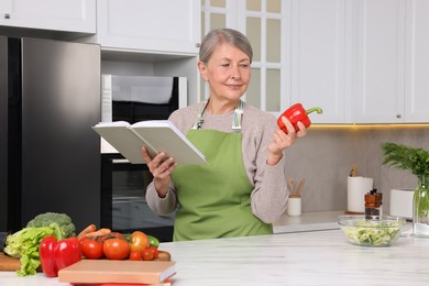 Happy woman with recipe book and bell pepper in kitchen