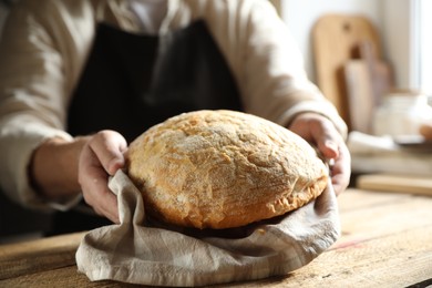 Man holding loaf of fresh bread at wooden table indoors, closeup