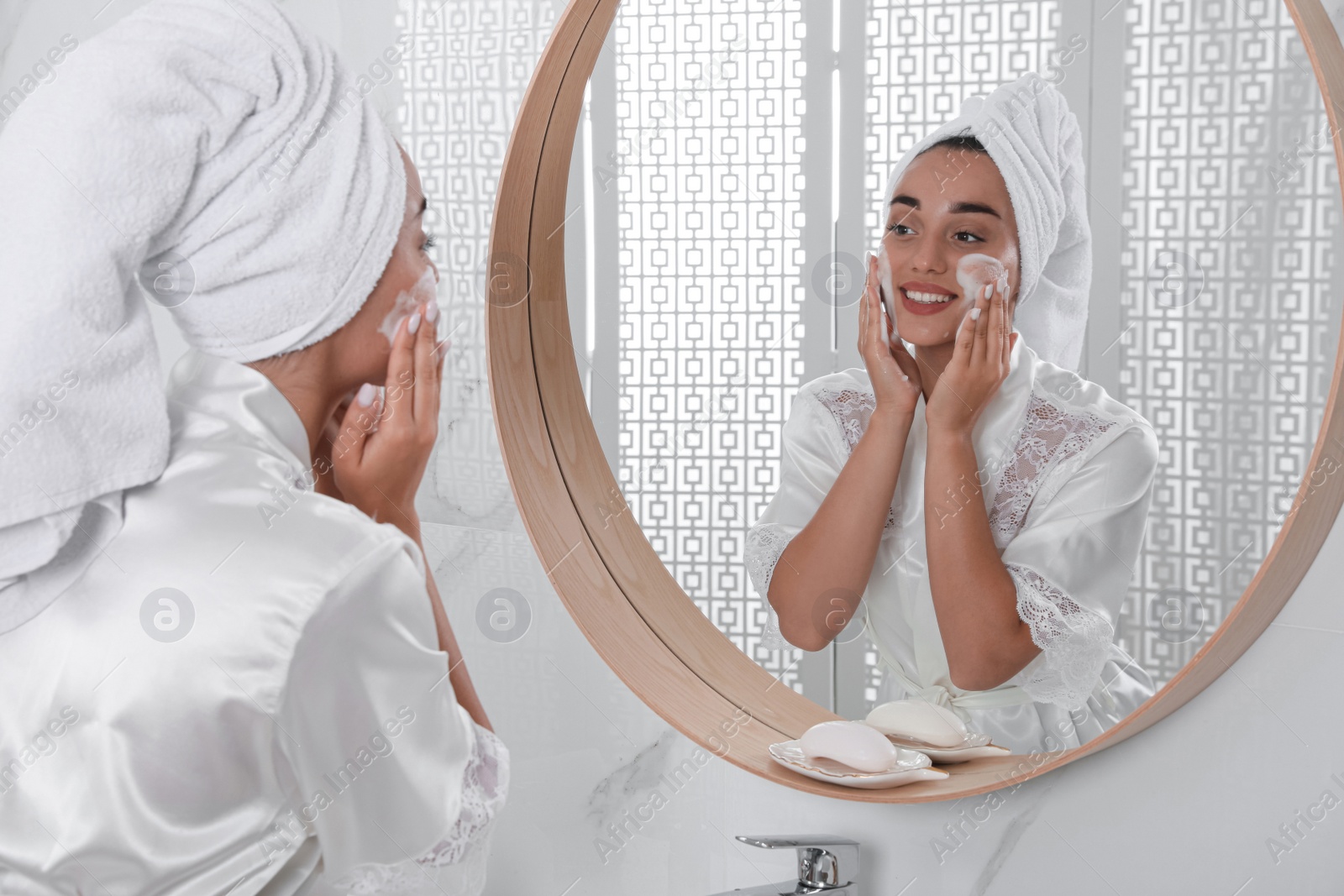 Photo of Beautiful young woman applying cleansing foam onto face near mirror in bathroom. Skin care cosmetic