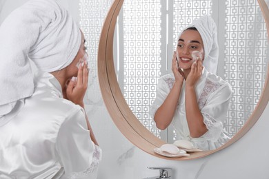 Beautiful young woman applying cleansing foam onto face near mirror in bathroom. Skin care cosmetic