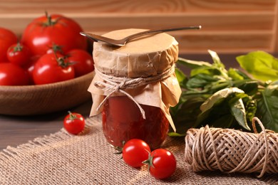 Jar of tasty tomato paste with spoon, ingredients and thread on wooden table