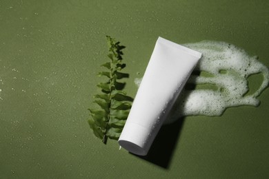 Cleansing foam, tube of cosmetic product and branch on wet dark green background, above view with space for text