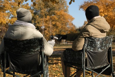 Couple with thermos sitting in camping chairs outdoors on autumn sunny day, back view