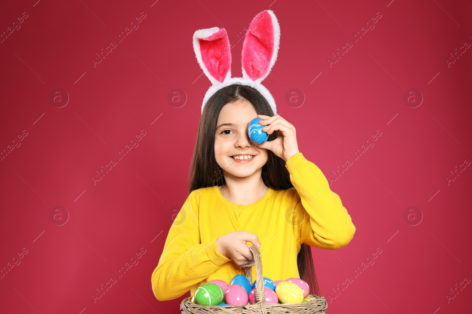 Photo of Little girl in bunny ears headband holding basket with Easter eggs on color background