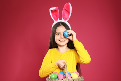 Little girl in bunny ears headband holding basket with Easter eggs on color background